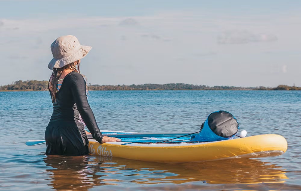 Girl in a gray hat standing on the water next to a surfboard and paddle