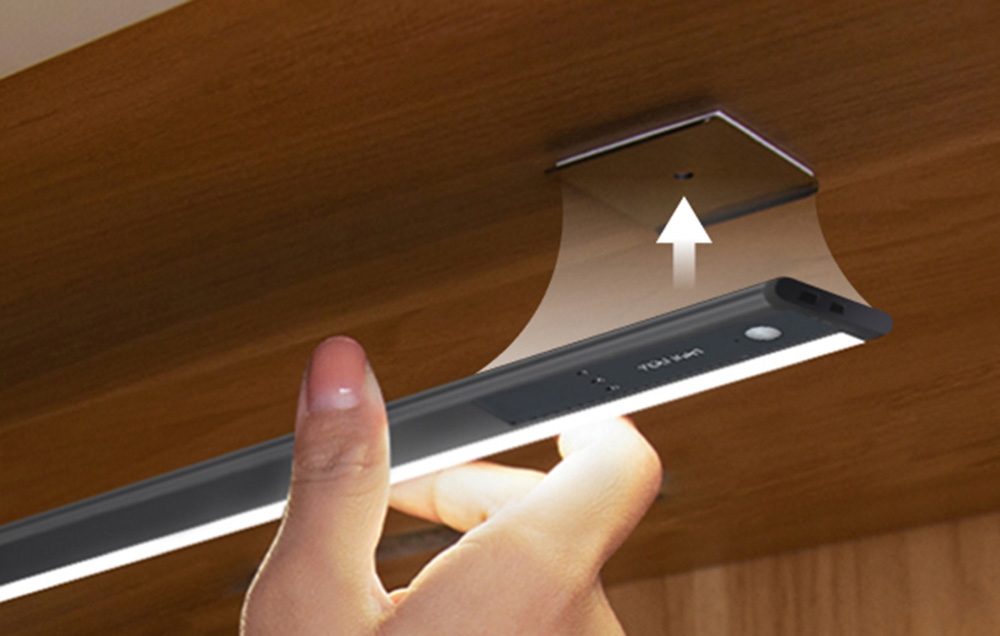 A hand mounting a Yeelight cabinet light under a wooden shelf using a magnetic plate.