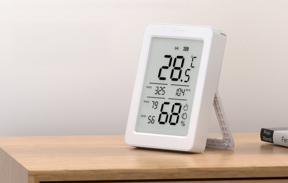 The Sonoff AirGuard TH sensor stands on a wooden shelf and displays the temperature and humidity on a large LCD screen.