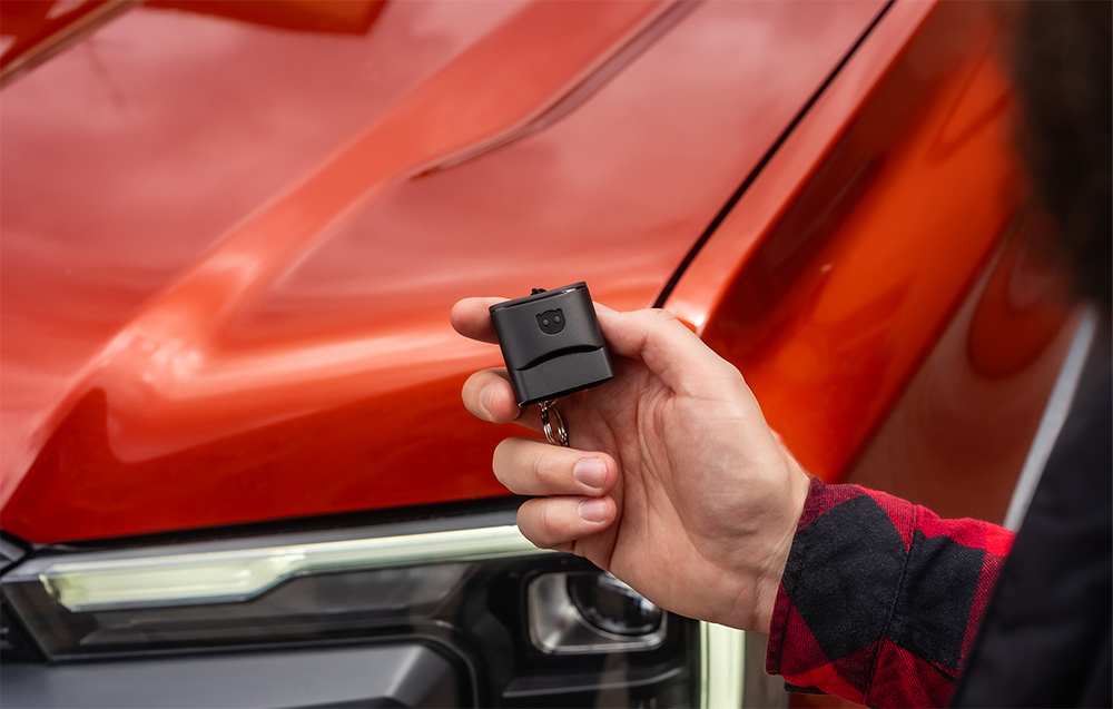 Person holding a black Carly diagnostic adapter against the open hood of an orange car