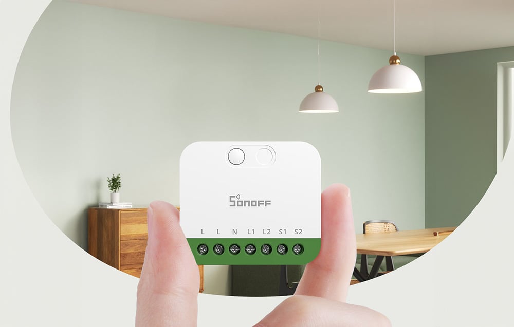 White Sonoff MINI-2GS smart switch held in hands, showing terminal with L, N, L1, L2, S1, S2 connection markings, against the background of a modern living room