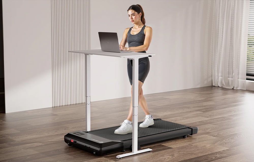 Woman working on a laptop at a white desk standing above the treadmill while walking