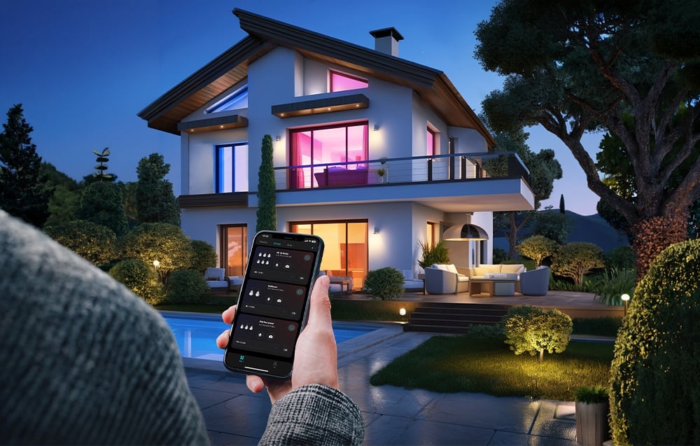 A hand holds a smartphone with a lighting control app open, set against the backdrop of a modern single-family home at dusk.