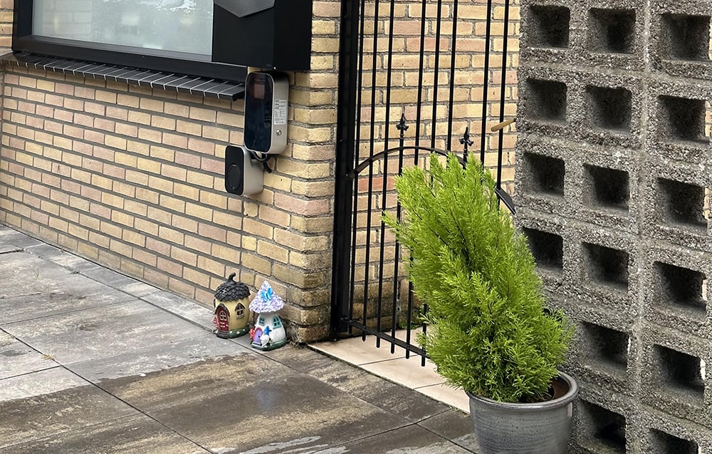 An electric car charging station mounted on the exterior wall of a building next to a gate and a planter with a plant.