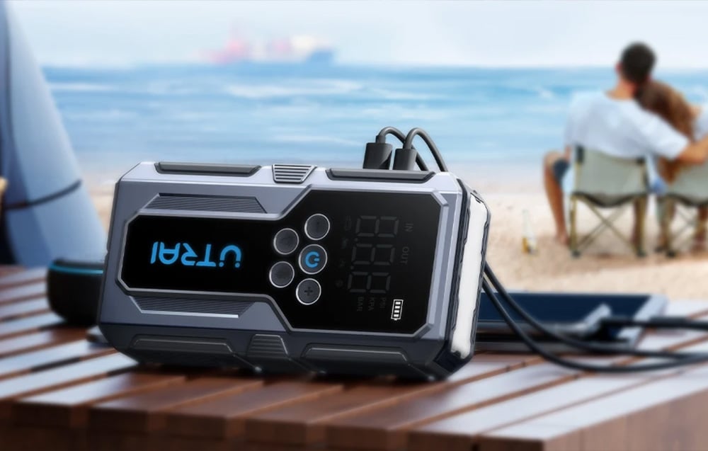 A car jump starter lies on a wooden table on the beach and is charging two mobile devices.