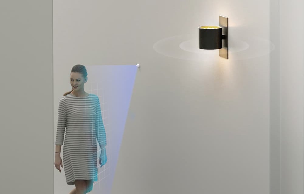 A woman walks past a wall-mounted motion detector with a visualization of the detection field and a lit lamp