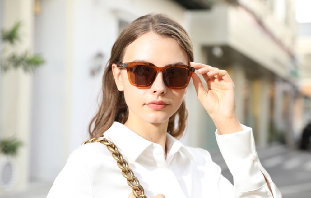 A woman in a white shirt adjusting her smart glasses with her hand, posing against a blurred background of a city street.