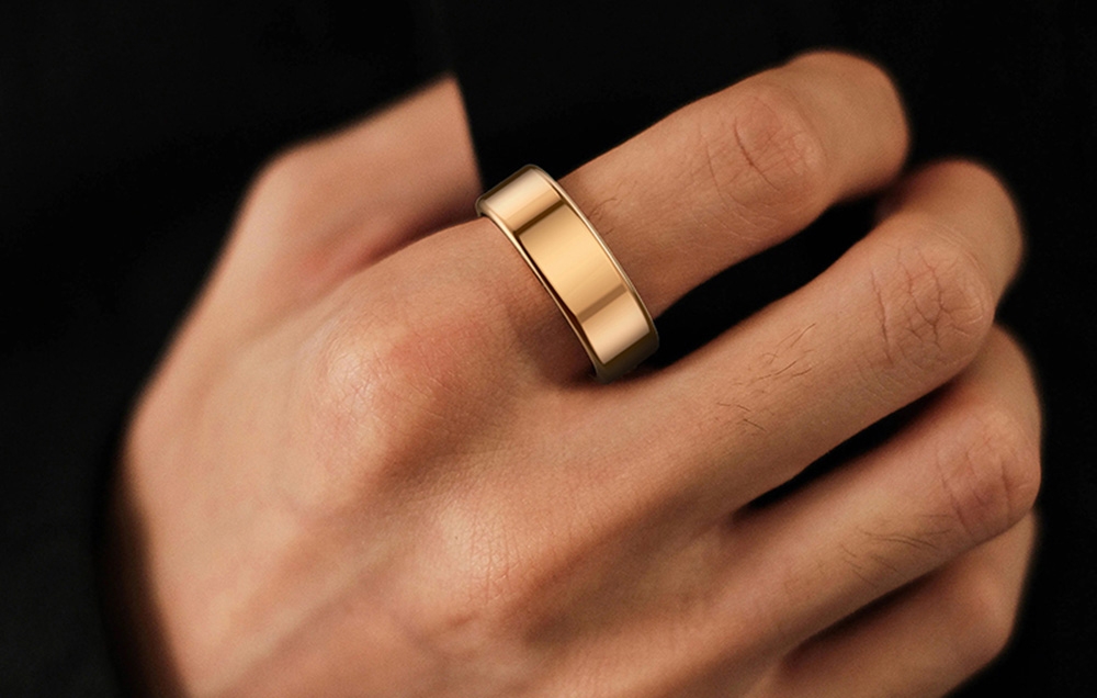 A man’s hand in a black jacket wearing a gold smart ring on his index finger