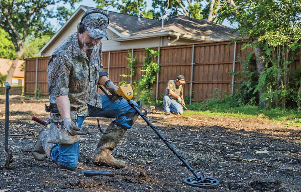 A man kneels and searches the ground with a metal detector in the backyard.