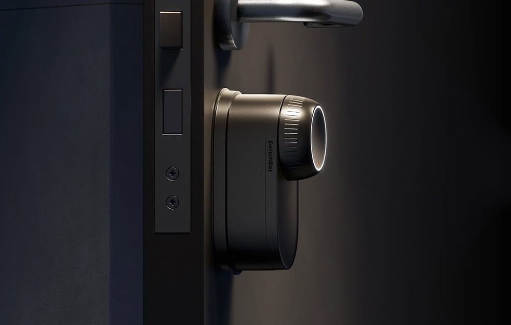 Black SwitchBot lock with logo mounted on dark door next to metal handle and lock cylinder