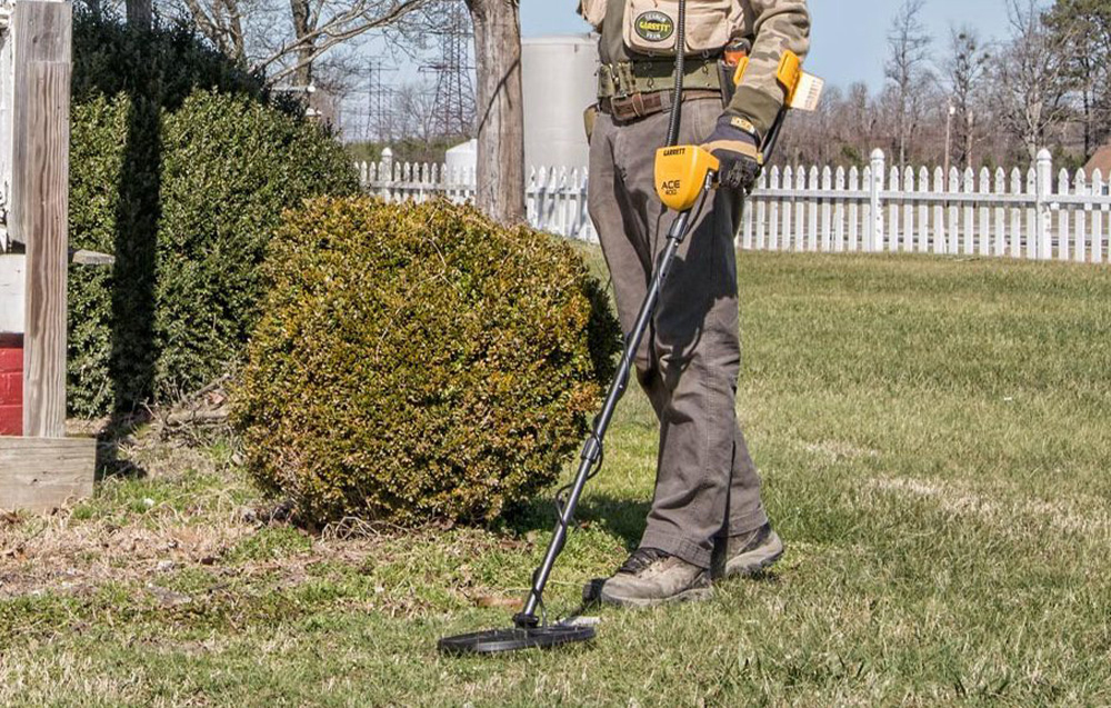 A person uses a metal detector on a lawn near a fence.