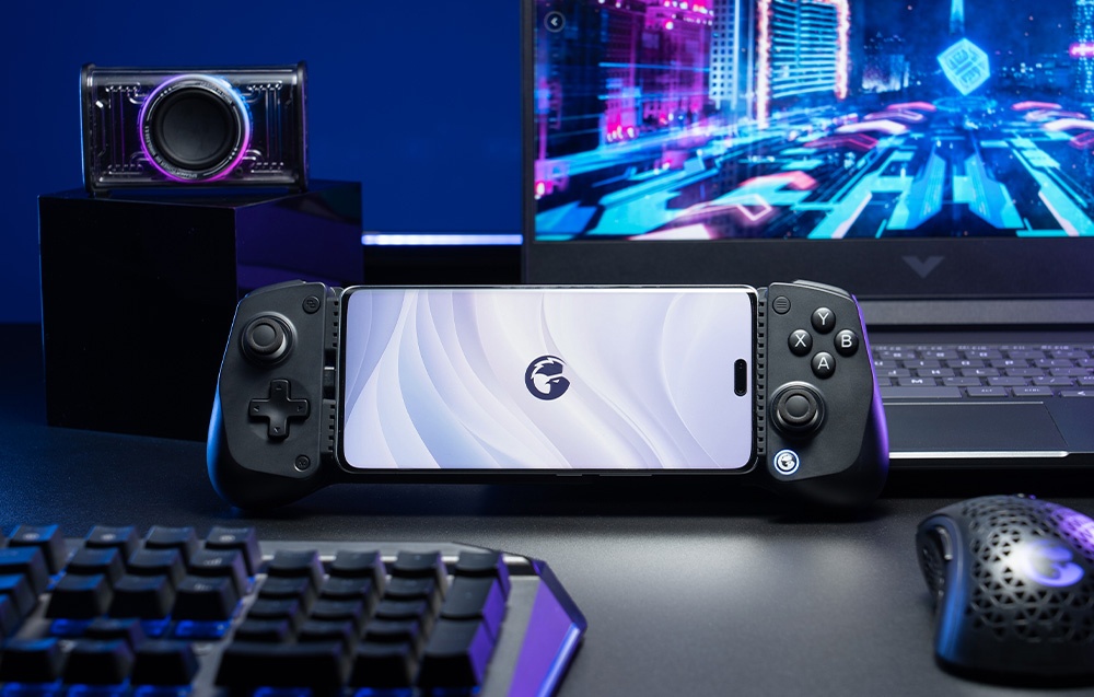 A smartphone mounted in a black GameSir controller set on a desk in front of a laptop and gaming accessories.