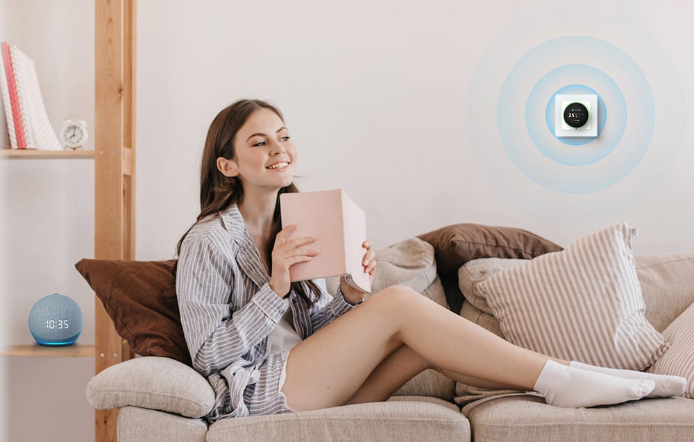 A smiling woman sits on the couch with a book. On the wall a smart thermostat