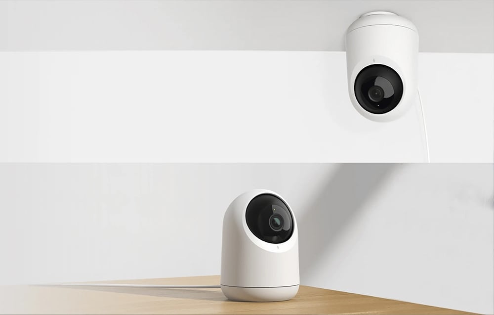 A white home security camera shown in two positions: standing on a light-colored table and mounted upside down on the ceiling.