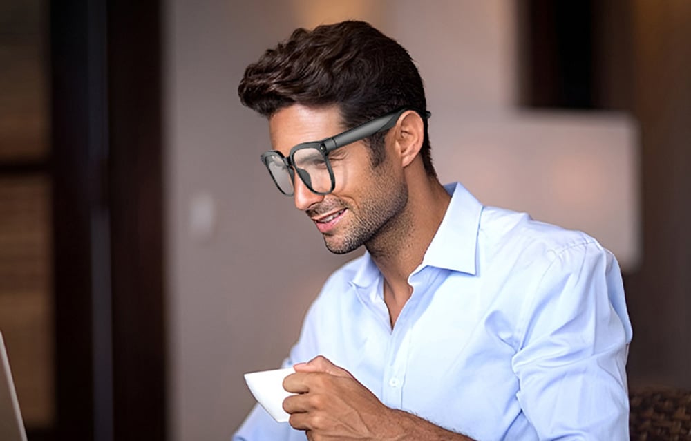 A smiling man in an elegant shirt drinks coffee and uses modern glasses while sitting at a computer. A woman in profile wearing glasses that direct sound direct