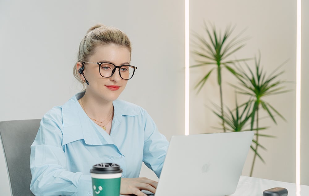 Woman in glasses working at a laptop, wearing black in-ear headphones with microphone installed