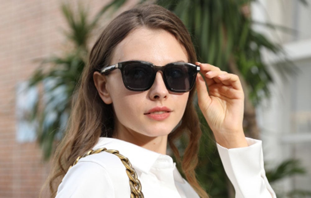 A woman in a white shirt and dark glasses, with buildings and plants in the background