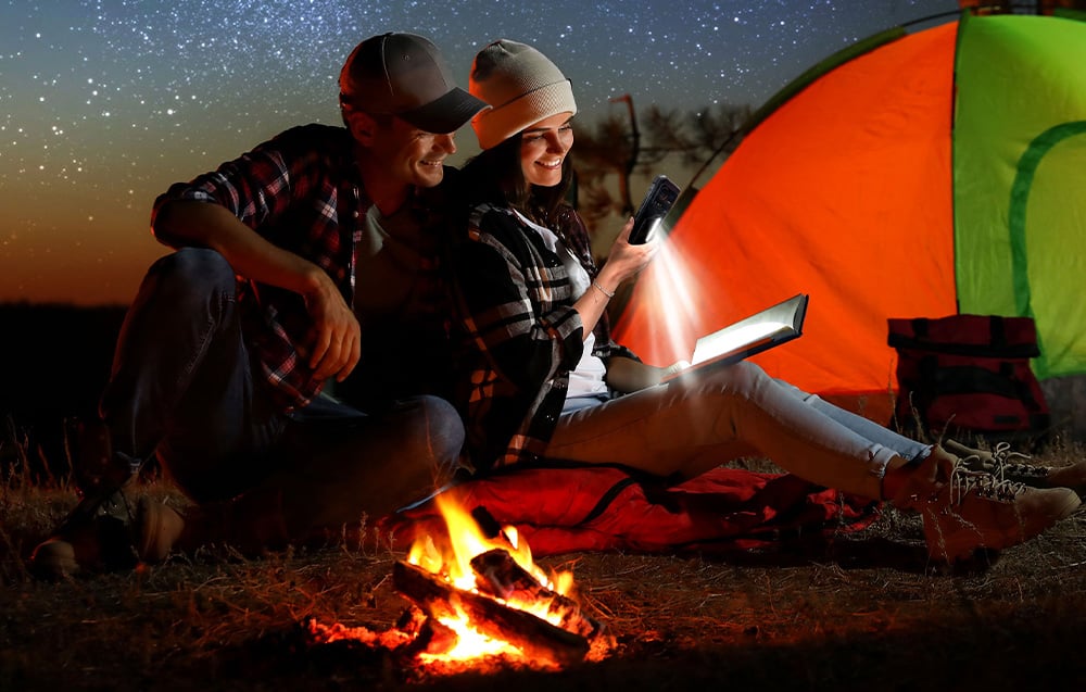A couple camping at night by a campfire under a starry sky; a woman illuminates the book she is reading with a powerful flashlight built into her smartphone.