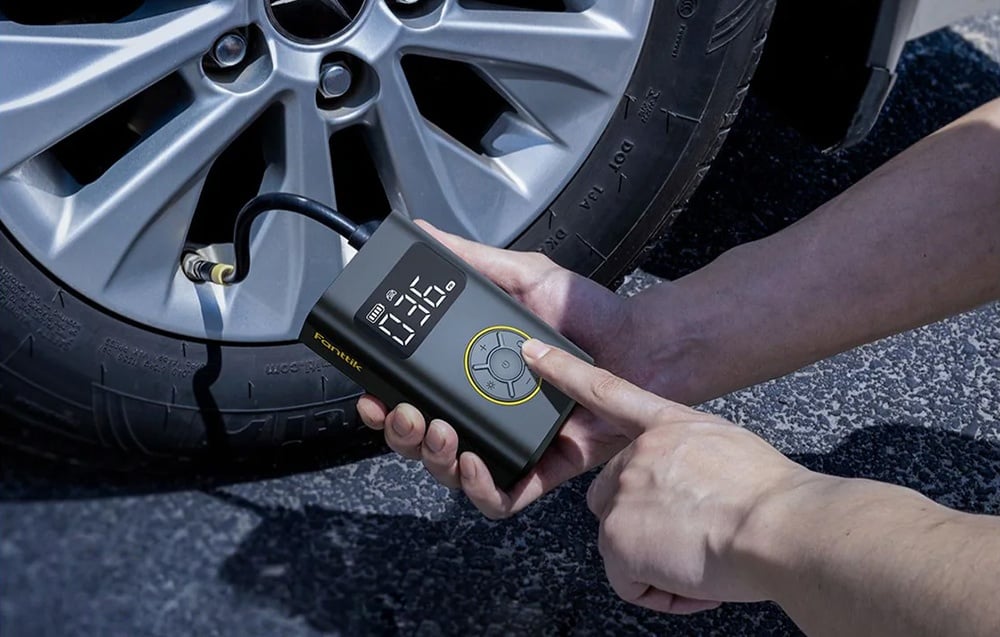 A man holds a mini compressor in his hands pumping up a car tire.