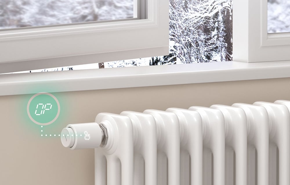 A radiator with a smart thermostatic head, an open window above it, and a winter landscape behind it