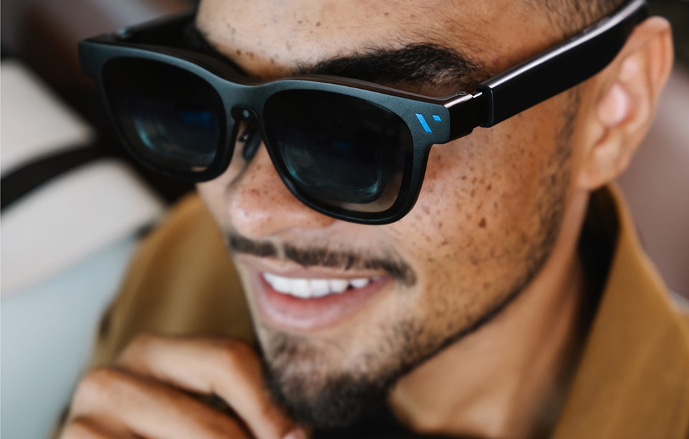 Close-up of the face of a man in black glasses with a blue accent, smiling and looking to the side
