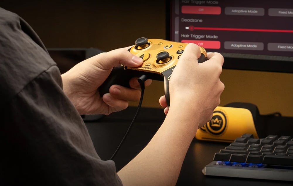 A person holding a wired game controller next to a desk with a monitor and keyboard