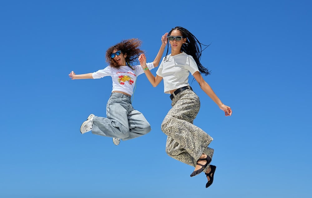 Two people wearing glasses are jumping in the air against a solid blue sky