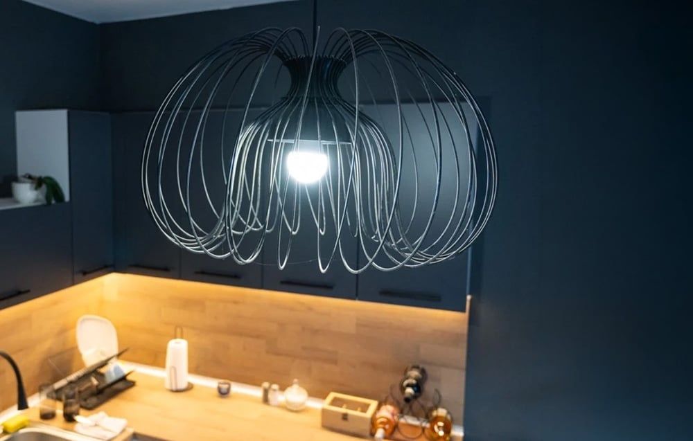 A large, black, wire chandelier with a warm-toned light bulb, hanging in a dark, modern kitchen with cabinets