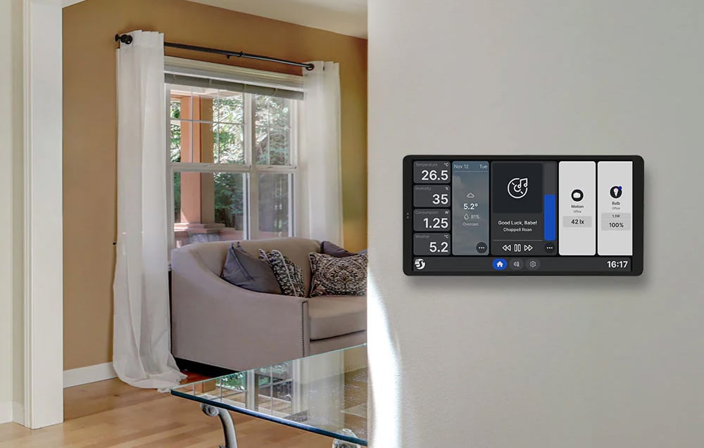 A Shelly X2i panel in a silver casing on the wall in the living room, displaying tiles with weather, temperature, and music player.