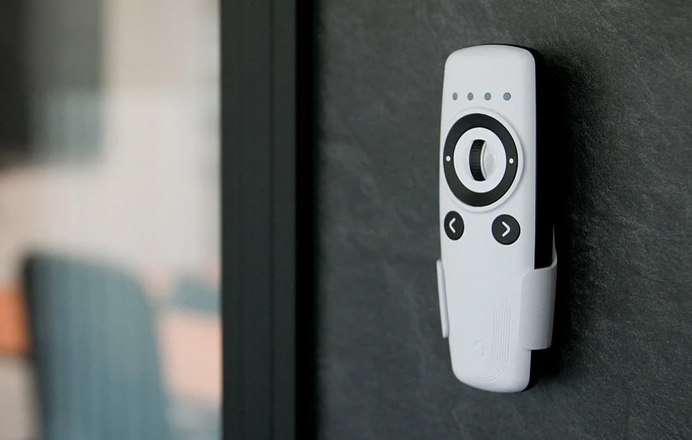 White Shelly remote control with knob and wall-mounted buttons, placed in a holder on a dark wall.