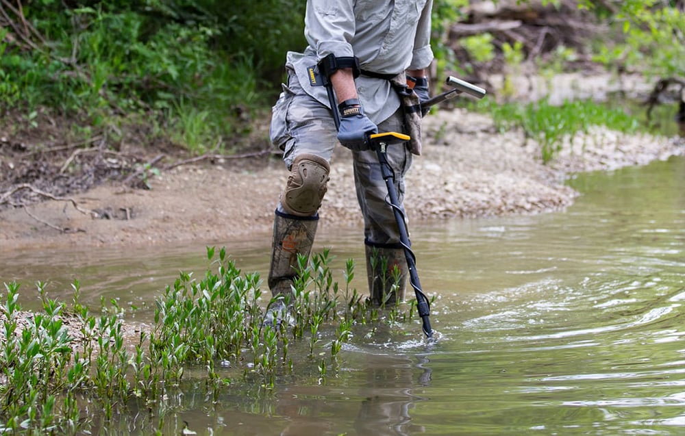 Person with metal detector searches shallow water near river bank.