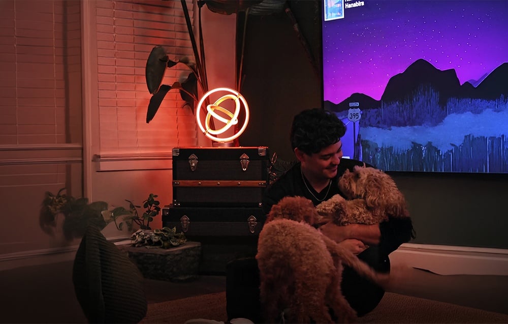 A man sitting on the floor with two dogs in a room with a TV and an orange-glowing desk lamp.