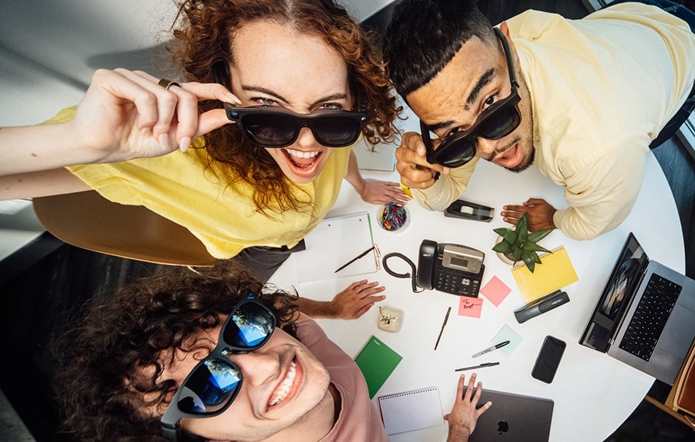 Three employees in XR glasses smile over a desk of laptops and sticky notes