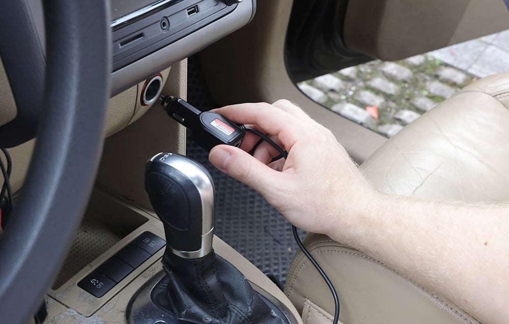 A man plugs a black power adapter into the cigarette lighter socket inside a brightly lit car