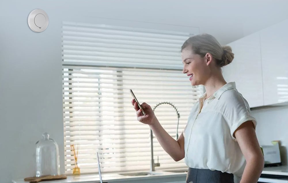 A smiling woman in a light-colored shirt checking her phone in a kitchen with a smoke detector visible on the wall