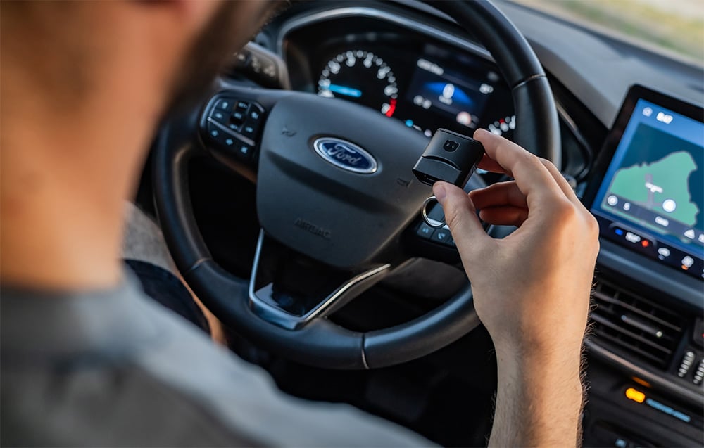 Driver holding Carly OBD2 adapter inside Ford car with media screen visible
