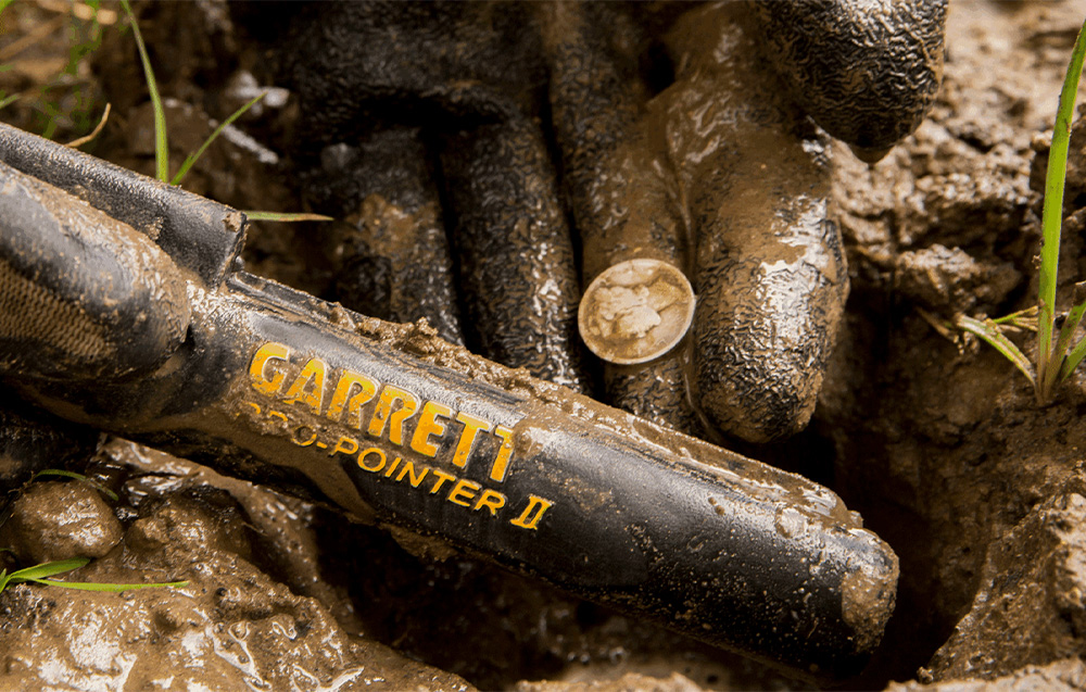 A gloved hand in the mud holds a coin next to a soiled metal detector.