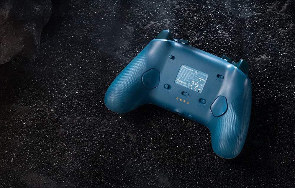 The back of the blue gaming controller with visible technical markings and textured grips, lying on a dark background