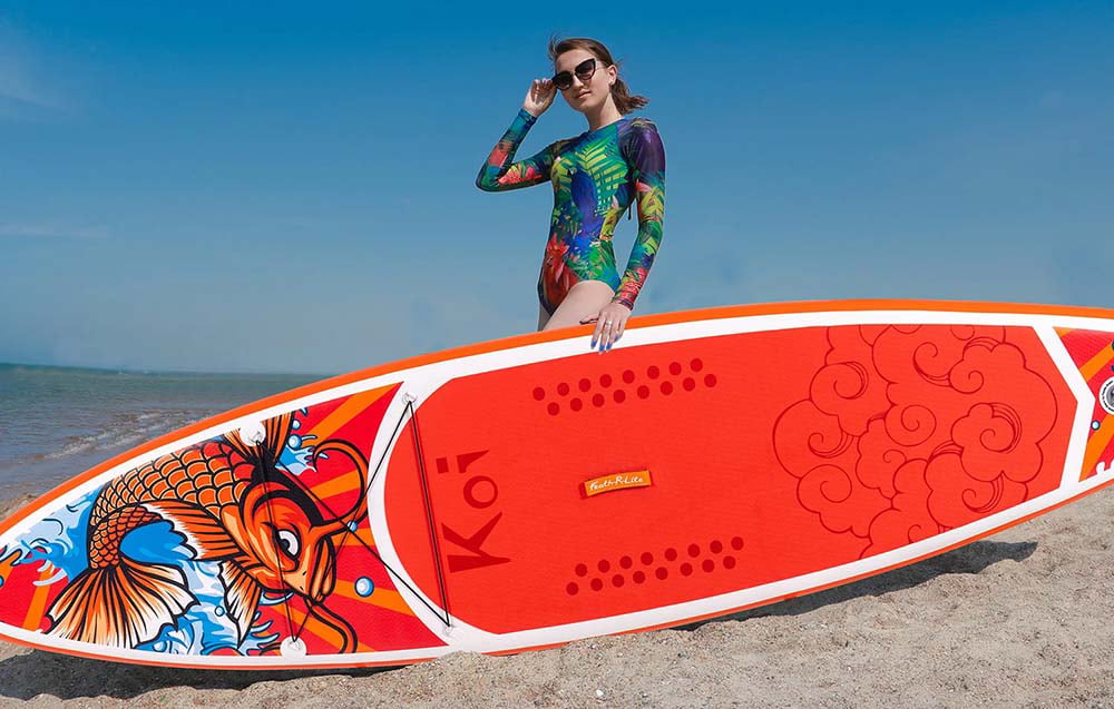 A woman in a colorful swimsuit and sunglasses holds a red surfboard with her hand while lying on the beach
