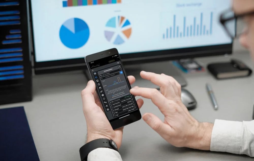 A man holding a smartphone displaying a graph of electricity consumption; a computer screen with diagrams is visible in the background.