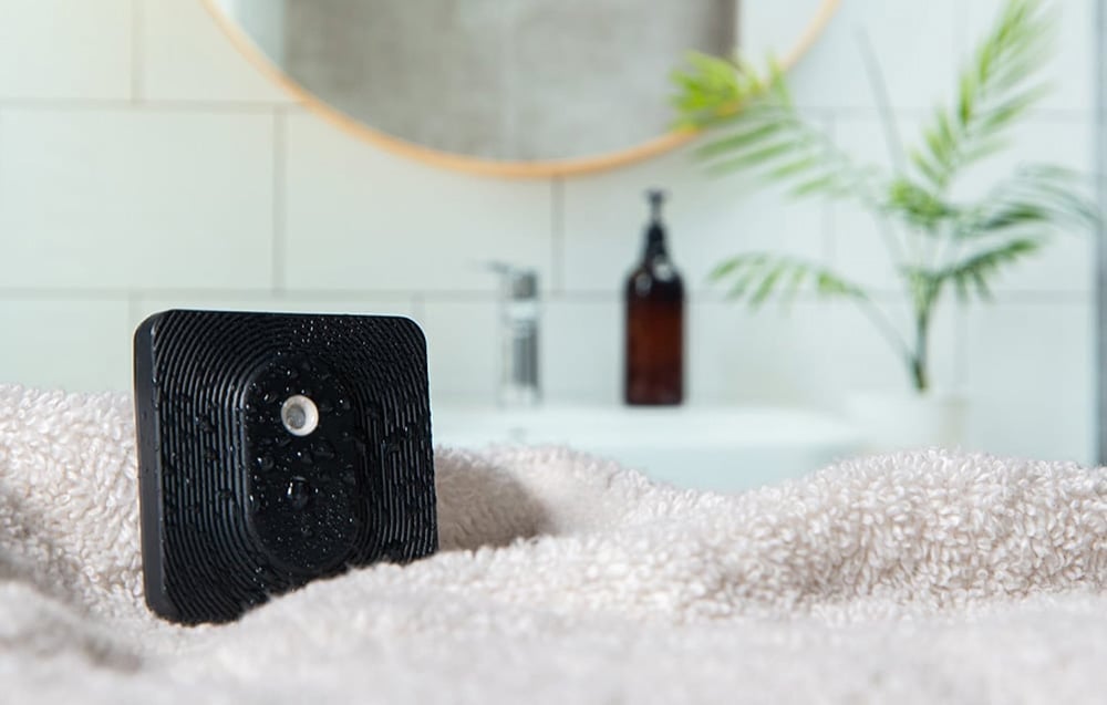 A black, damp Shelly sensor placed on a white towel in a bright bathroom with a mirror in the background