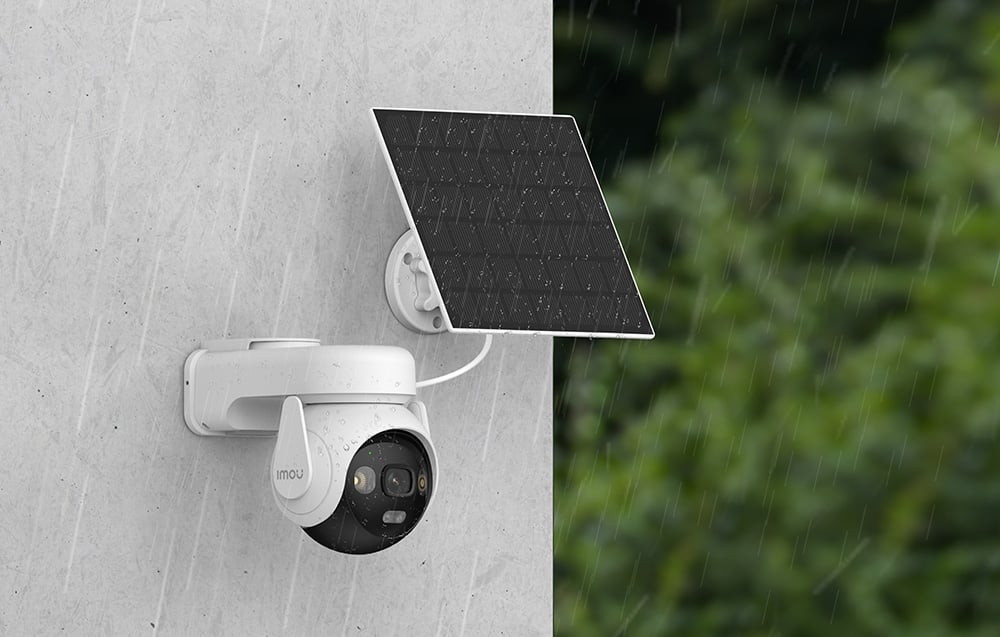 White pan-tilt camera and solar panel mounted on a gray wall in the rain, with green trees in the background