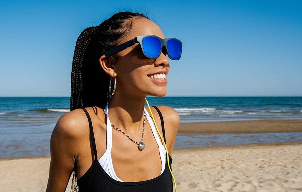 A woman wearing glasses with blue lenses and a black top is standing on a sandy beach by the sea
