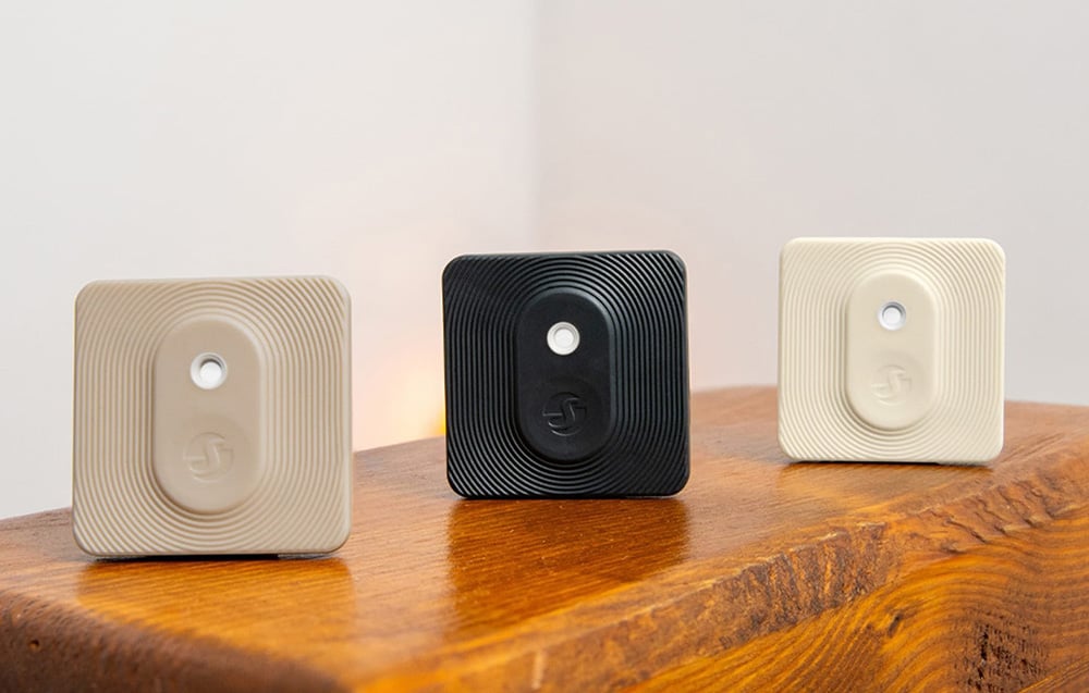 Three square Shelly sensors in beige, black, and cream colors arranged on a wooden base
