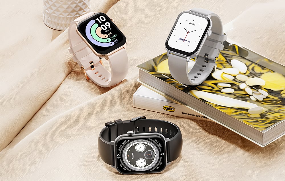 Three Colmi smartwatches (pink, gray, black) in an elegant arrangement on fabric next to books