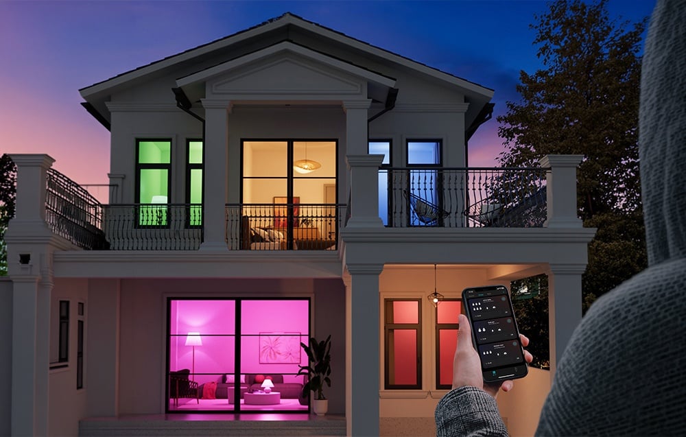 A two-story house at night, with windows lit up in green, pink, and blue, and a hand holding a smartphone with the app open.