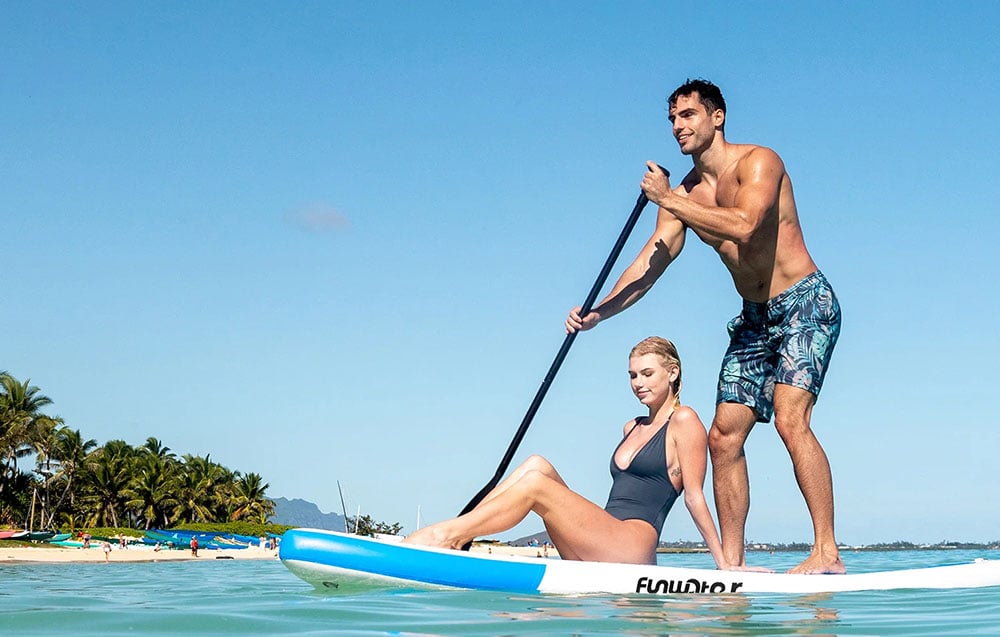 A man stands on a Funwater surfboard and paddles, with a woman sitting next to him