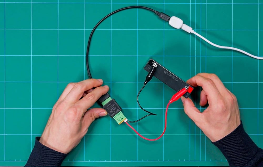 Two hands holding a black module with a green connector and a battery with the number F232243224, connected by cables above the mat