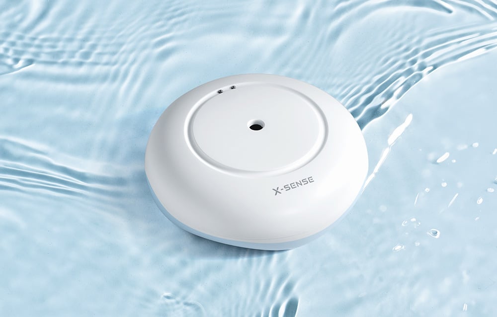 A round, white X-Sense flood sensor floating on the surface of rippling water