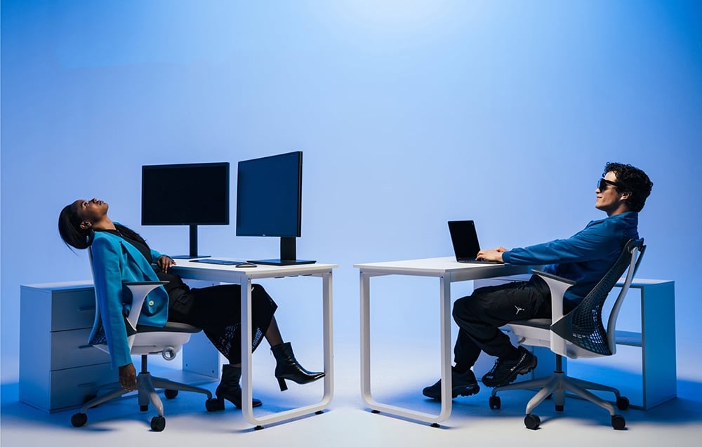 Two people are working at desks against a blue background. The man is wearing modern glasses.